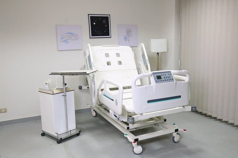 Santron Pro ICU Bed, backrest raised, in a styled patient room with overbed table and cabinet