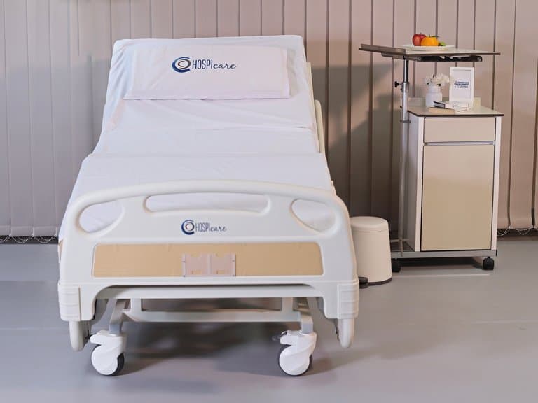 HOSPIcare hospital bed made up with branded linen in a furnished patient room with overbed table and bedside cabinet