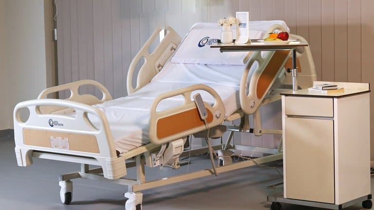 غرفة مريض مجهزة بسرير هوسبي كير للأوزان الثقيلة مفروش بأغطية تحمل الشعار مع طاولة فوق السرير وكومودينو — MLP-11