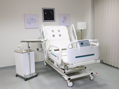 Santron Pro ICU Bed, backrest raised, in a styled patient room with overbed table and cabinet