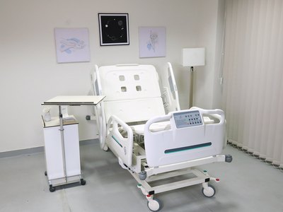 Santron Hospital Bed, backrest raised, in a styled patient room with overbed table and cabinet